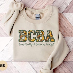 Puede incluir: Una sudadera de color arena con el texto "BCBA Board Certified Behavior Analyst" en letras verdes con un diseño floral.