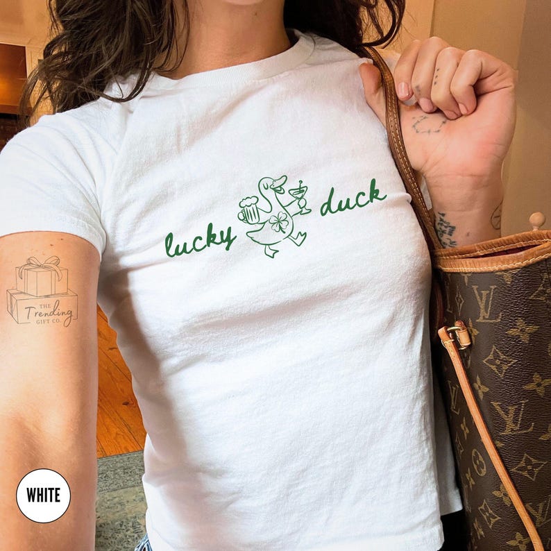 Op de afbeelding: Wit T-shirt met een groene afbeelding van een eend met een bier en een cocktail, met de woorden "lucky duck" in groene letters. Het shirt heeft korte mouwen en een ronde hals.