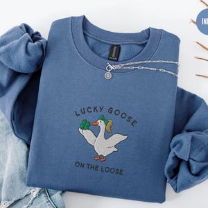 May include: Indigo blue sweatshirt with a white goose wearing a green shamrock hat and holding a shamrock. The text "Lucky Goose on the Loose" is embroidered on the sweatshirt.