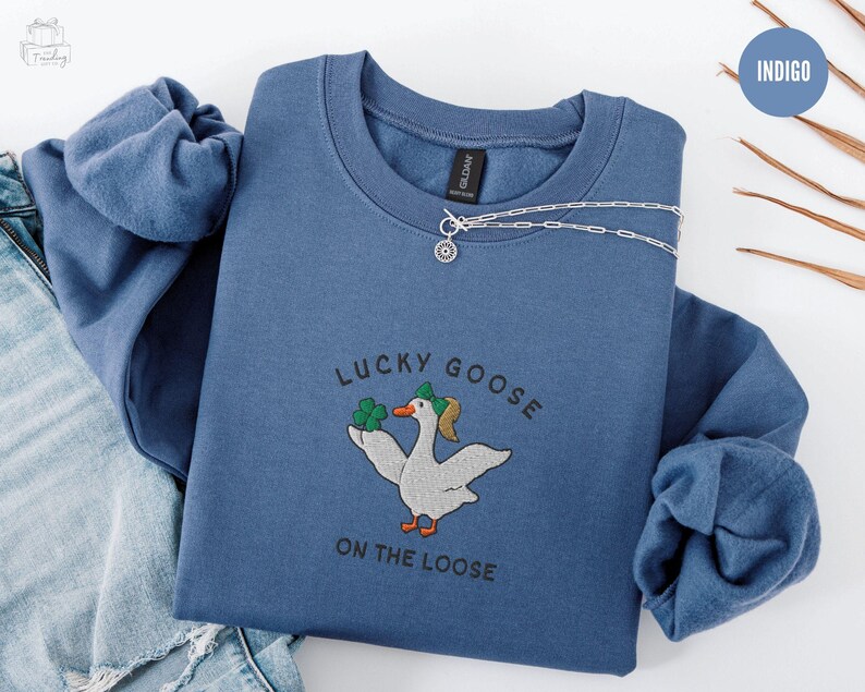 May include: Indigo blue sweatshirt with a white goose wearing a green shamrock hat and holding a shamrock. The text "Lucky Goose on the Loose" is embroidered on the sweatshirt.