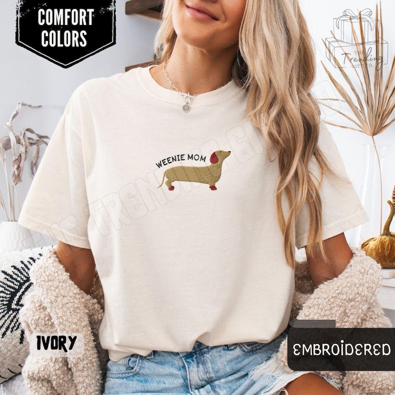 May include: Ivory-colored t-shirt with the text "Weenie Mom" and an embroidered dachshund dog. The shirt also has the words "Comfort Colors" and "Embroidered" printed on it.