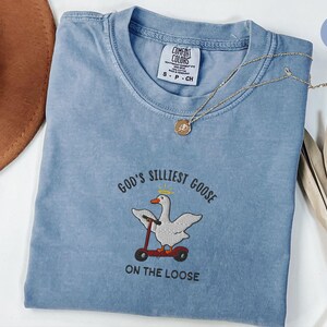 May include: A blue jean t-shirt with a white embroidered goose riding a scooter. The text "God's Silliest Goose On The Loose" is embroidered on the shirt.