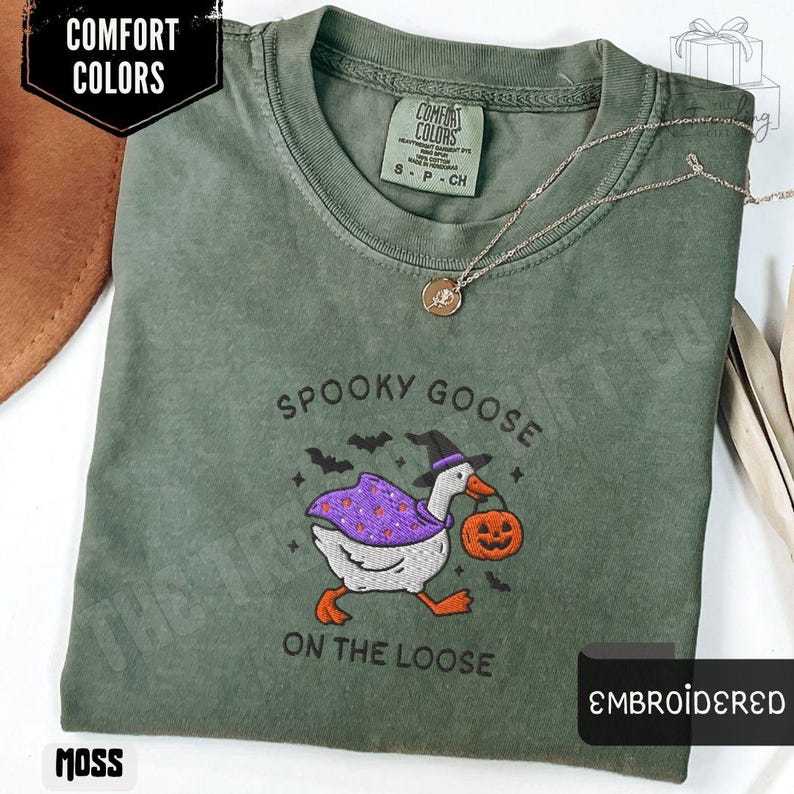 May include: Moss green t-shirt with an embroidered design featuring a goose in a witch hat and cape, carrying a jack-o'-lantern. The text "SPOOKY GOOSE ON THE LOOSE" is above and below the goose. The shirt has a crew neck and short sleeves.