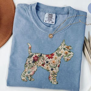 Puede incluir: Una camiseta azul claro con una silueta de perro bordada con flores. La camiseta tiene una etiqueta que dice "Comfort Colors" y "S-P-CH".