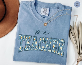 May include: A blue jean Comfort Colors t-shirt with the words "p.e. TEACHER" in a floral design. The shirt has a crew neck and short sleeves. A gold necklace with a pendant is draped across the shirt.