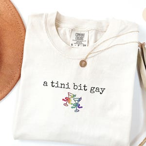May include: Ivory t-shirt with the text "a tini bit gay" and a rainbow design of martini glasses.
