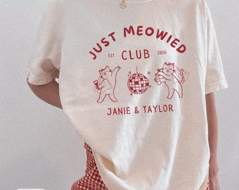 May include: Ivory-colored t-shirt with a red graphic that reads "Just Meowied Club" with the year 2026. The graphic features two cartoon cats, a disco ball, and the names "Janie & Taylor".