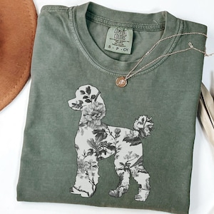 May include: A moss green t-shirt with a floral poodle silhouette design. The shirt has a tag that reads "Comfort Colors" and "S - P - CH".