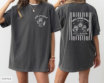 Może przedstawiać: Ciemnoszary t-shirt z krótkim rękawem z białą grafiką szopa pracza w koronie na piersi z napisem "girls gone feral". Z tyłu znajduje się większa grafika z dwoma szopami, kulą dyskotekową i napisem "GIRLS GONE FERAL est. 2026 Lauren's Bachelorette MIAMI BEACH FL" w ramce z pionowych pasków.