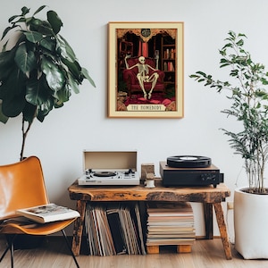 May include: A framed print featuring a skeleton in a red armchair, holding a glass. The print is labeled "THE HOMEBODY" and is displayed above a wooden table with a record player and vinyl records. The room has a vintage aesthetic.