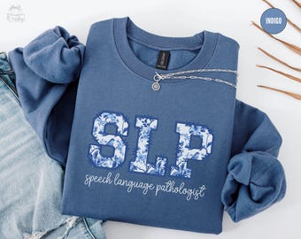 May include: Indigo blue sweatshirt with the letters "SLP" in a floral pattern, with the words "speech language pathologist" below. A silver necklace with a circular pendant is on the sweatshirt.