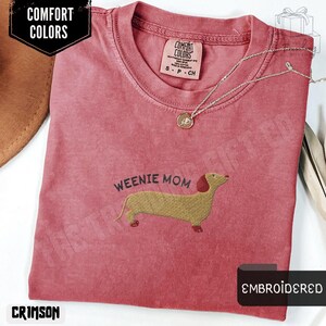 May include: Crimson-colored t-shirt with the text "Weenie Mom" and an embroidered dachshund. The shirt has a crew neck and short sleeves. A silver necklace with a pendant is draped across the shirt. The shirt is labeled "Comfort Colors".