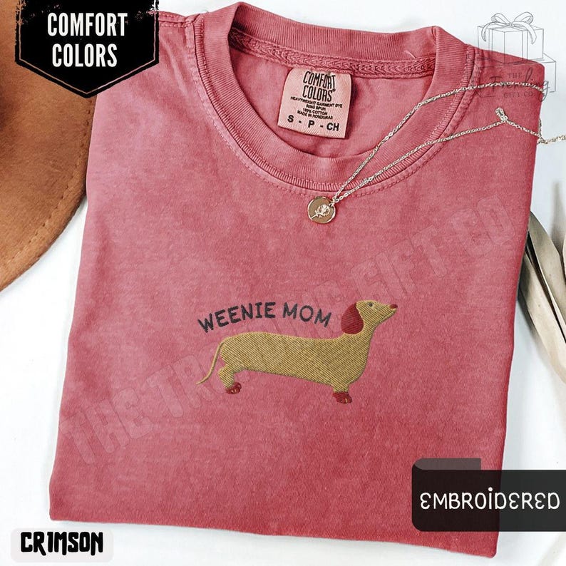 May include: Crimson-colored t-shirt with the text "Weenie Mom" and an embroidered dachshund. The shirt has a crew neck and short sleeves. A silver necklace with a pendant is draped across the shirt. The shirt is labeled "Comfort Colors".