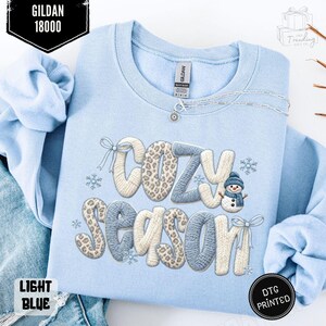 Peut inclure: Sweat-shirt bleu clair avec l'inscription "COZY SEASON" dans un design texturé, avec un bonhomme de neige et des flocons de neige. Le sweat-shirt a un col rond et des manches longues. La marque "GILDAN" est visible.