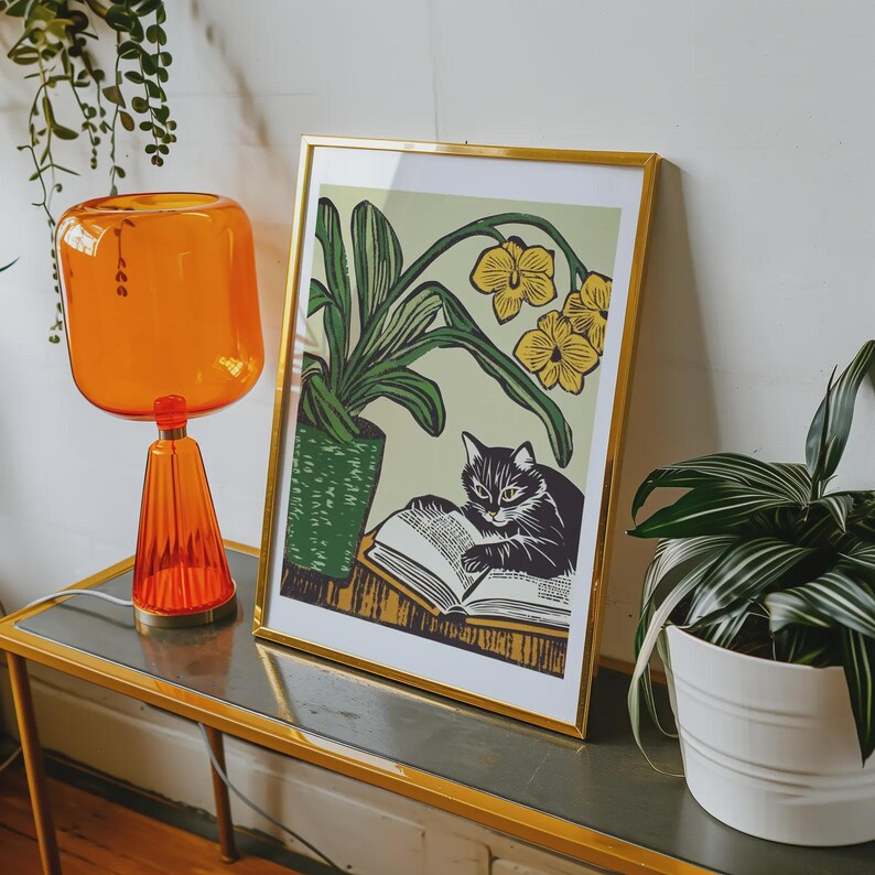 May include: A framed print of a black cat reading a book with a yellow orchid plant in a green pot. The print is in a gold frame and is sitting on a table with a glass top.