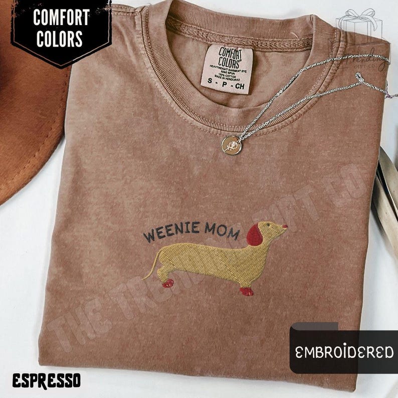 May include: Espresso-colored t-shirt with an embroidered dachshund design and the text "Weenie Mom." The shirt features a crew neck and short sleeves. A silver necklace with a pendant rests on the shirt. The shirt is labeled "Comfort Colors."