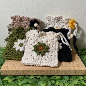 May include: A collection of handmade crochet pouches in various colors, including green, cream, black, and brown. Each pouch features a crocheted flower design in contrasting colors. The pouches are displayed on a wooden surface.