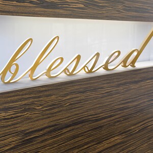 May include: A gold-coloured 'blessed' sign in cursive script. The word is displayed on a white shelf, set against a backdrop of dark wood grain. The sign is a decorative item, suitable for home decor.