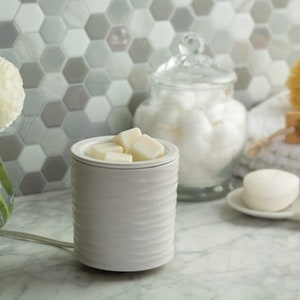 Könnte beinhalten: Ein weißer Keramik-Duftwachsverdampfer mit einem strukturierten, geriffelten Design, gefüllt mit quadratischen Wachs-Melts. Ein Glasbehälter mit Wattebällchen und ein Stück Seife befinden sich im Hintergrund, was auf ein Spa- oder Badezimmerumfeld hindeutet.