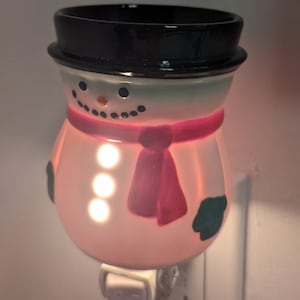 Könnte beinhalten: Ein schneemannförmiges Nachtlicht mit schwarzem Hut, rotem Schal und grünen Fäustlingen. Das Nachtlicht hat ein lächelndes Gesicht und drei leuchtende Lichter. Es ist an eine Steckdose angeschlossen.