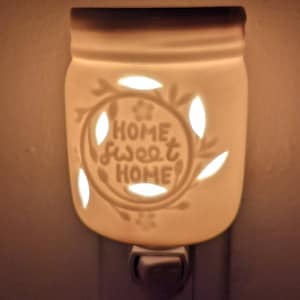 Könnte beinhalten: Ein weißes Keramik-Nachtlicht in Form eines Einmachglases. Es hat ausgeschnittene Blatt- und Blumenmuster mit dem Text "HOME sweet HOME". Das Nachtlicht wird direkt in eine Steckdose gesteckt.
