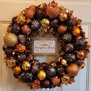 May include: A festive autumn wreath with a variety of gold, bronze, and brown ornaments and faux leaves. A small wooden sign in the center reads "Autumn Acres Pumpkin Patch" with text about apple cider, hay rides, and hot cocoa. The wreath is a seasonal decoration.