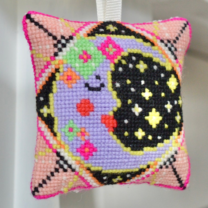 May include: A square, cross-stitched ornament featuring a smiling crescent moon and stars against a black background. The moon is lavender with colorful floral accents. The ornament has a pink border and a white ribbon for hanging.