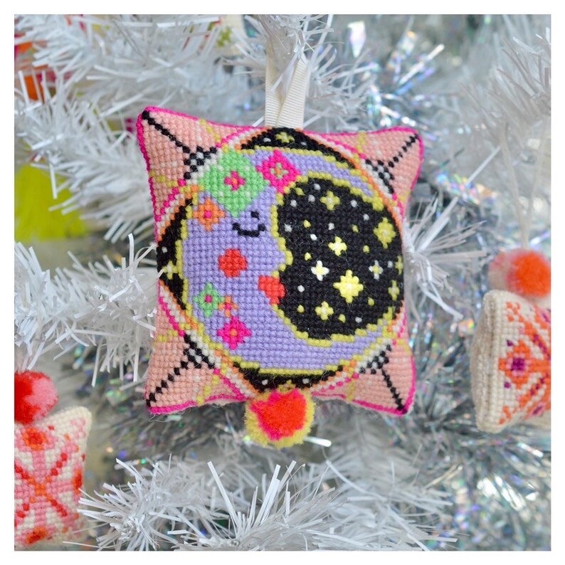 May include: A decorative, cross-stitched Christmas ornament in the shape of a pillow. The ornament features a smiling moon with stars on a black background, surrounded by a pink and black border. A small orange pom-pom hangs from the bottom.