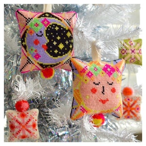 May include: Handmade star-shaped Christmas ornaments with cross-stitch designs. One features a crescent moon and stars, another a cat face with floral accents. Additional ornaments with geometric patterns are also visible, all hanging on a white Christmas tree.