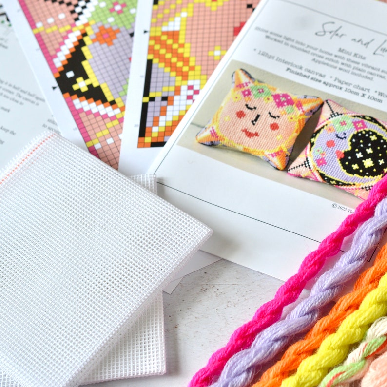 May include: A craft kit with a cross-stitch pattern for a pillow. The kit includes a white mesh canvas, colorful embroidery floss, and printed instructions. The instructions show a finished pillow with a face and floral design. The text on the instructions reads "Solar and Luna Mini Kite".