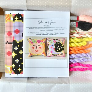 May include: A craft kit with the text "Solar and Lunar Mini Kits" includes a paper chart, wool, and needle. The kit features two cross-stitch pillow designs: a sun and a moon. The finished size is approximately 4 inches x 4 inches.
