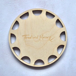 Puede incluir: Un portahilos de madera clara de forma circular, con una serie de cortes semicirculares en el borde. El texto "Thread and Mercury" está grabado en la madera. Diseñado para organizar carretes de hilo.