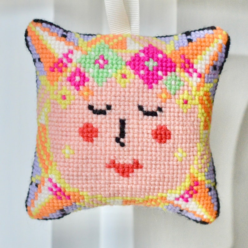 May include: A square, cross-stitched ornament featuring a smiling face with closed eyes. The face is primarily pink with red cheeks and a red mouth. The ornament has a floral crown and a colorful border with orange, yellow, and purple accents. A white ribbon is attached for hanging.