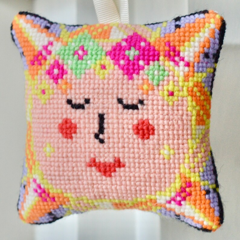 May include: A square, cross-stitched ornament featuring a face with closed eyes, rosy cheeks, and a red mouth. The ornament has a peach background with a colorful border of orange, yellow, green, and purple. A cream ribbon is attached for hanging.