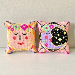 May include: Two small, square, handmade pillows. One features a face with closed eyes and a floral crown, in shades of pink, orange, and yellow. The other pillow has a crescent moon and stars in purple, black, and pink.