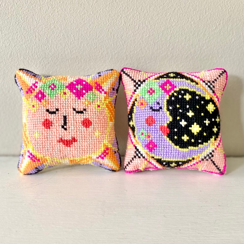 May include: Two small, square, handmade pillows. One features a face with closed eyes and a floral crown, in shades of pink, orange, and yellow. The other pillow has a crescent moon and stars in purple, black, and pink.