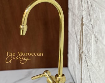 May include: A gold-colored kitchen faucet with a curved spout and lever handle. The faucet is set against a white and gray countertop and a textured white wall. The Moroccan Gallery logo is visible.