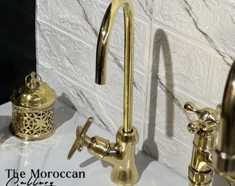 May include: A gold-colored faucet with a curved spout and cross-shaped handles. The faucet is part of a bathroom fixture set. The image also shows a decorative lantern and the text "The Moroccan Gallery".