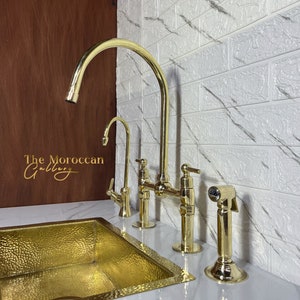 Könnte beinhalten: Ein goldfarbener Küchenarmaturen-Set mit passendem Waschbecken. Der Wasserhahn hat einen hohen Auslauf und einen Seitensprüher. Das Waschbecken ist rechteckig mit gehammerter Textur. Das Logo The Moroccan Gallery ist sichtbar.