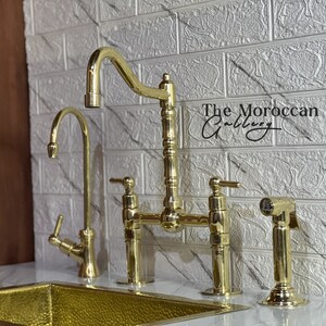 May include: A gold-colored kitchen faucet set with a bridge faucet, a separate water spout, and a side sprayer. The faucet set is displayed against a white brick-patterned wall. The Moroccan Gallery is written on the wall.