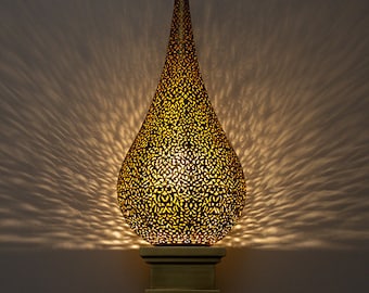May include: A gold-colored, teardrop-shaped wall sconce with intricate cut-out patterns. The sconce is mounted on a square base and emits a warm, inviting light, creating a decorative lighting effect. The design is reminiscent of Moroccan or Middle Eastern decor.