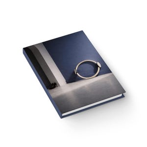 Könnte beinhalten: Ein Hardcover-Buch mit blauem Einband, das ein silbernes Armband zeigt. Der Buchumschlag zeigt ein Foto eines modernen Innenraums mit einer blauen Wand und einem grauen Boden. Das Armband ist oval mit zwei silbernen Enden.