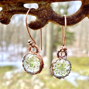 May include: A pair of handcrafted copper earrings. Each earring features a clear, round glass orb encasing a delicate green and white floral arrangement. The earrings hang from copper ear wires.