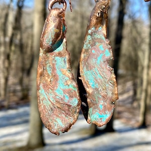 May include: A pair of handcrafted copper earrings with a unique, organic design. The earrings feature a teardrop shape with a textured, weathered appearance. The copper has a natural patina with shades of brown and turquoise, creating a rustic, artistic look.