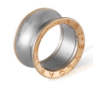May include: A two-tone ring with a silver-colored center and a gold-colored outer band. The gold band features engraved lettering. The ring has a modern, minimalist design, suitable for everyday wear or special occasions. The ring is a piece of jewelry.