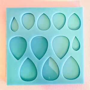 Puede incluir: Un molde cuadrado de silicona azul claro con nueve cavidades en forma de lágrima de varios tamaños. El molde está diseñado para manualidades, posiblemente para resina u otros materiales, y es adecuado para crear pendientes o colgantes.