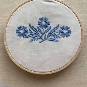 May include: A white embroidery hoop with a blue floral design embroidered on the fabric.