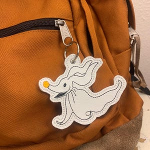 May include: White felt keychain with black stitching in the shape of Zero from the Nightmare Before Christmas. Zero has a yellow circle nose and is attached to a silver key ring.