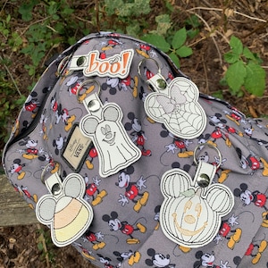 May include: Five Halloween-themed Mickey Mouse keychains with different designs, including a ghost, a pumpkin, a spiderweb, a candy cane, and a "boo!" keychain. The keychains are attached to a gray backpack with a Mickey Mouse pattern.