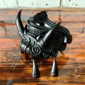 Puede incluir: Una máscara de plástico negra, detallada, con un diseño de cuernos. La máscara tiene una superficie texturizada y detalles intrincados. La máscara es de estilo japonés tradicional.